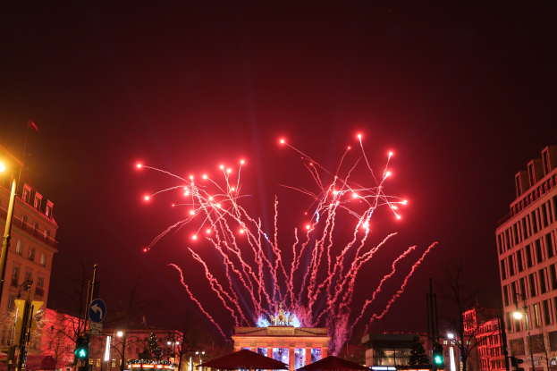 Eine belebte Stadtstraße an einem Silvesterabend in Berlin mit Gebäuden, Bäumen, Laternen, Ampeln, Schildern, Zelten, Menschen und einem prächtigen Feuerwerk am Himmel.