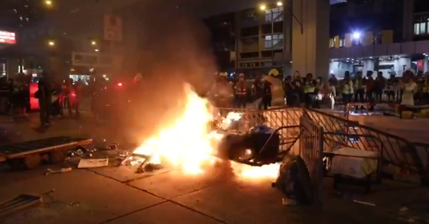 Eine Gruppe von Menschen steht um ein Feuer in der Mitte einer Straße, umgeben von Barrikaden und anderen Gegenständen, mit Gebäuden, Laternenmasten und einer Tafel mit Text im Hintergrund unter einem Nachthimmel.