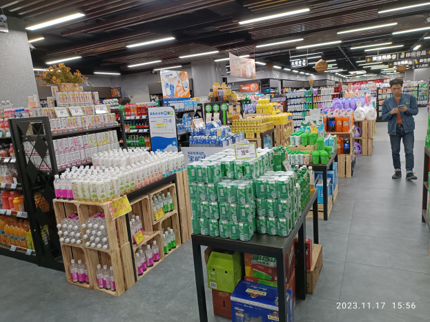 Ein Mann steht vor einem chinesischen Supermarkt mit verschiedenen Getränken, darunter Flaschen und Kartons, die auf Regalen angeordnet sind, mit Texttafeln und Deckenleuchten sowie einem Wasserzeichen am unteren Bildrand.