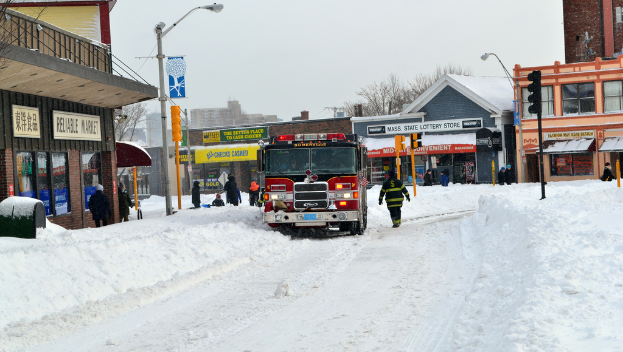 Eine Feuerwehr ist vor Gebäuden im Schnee geparkt, mit Geschäften und Laternenmasten links, einer Ampel in der Mitte und Schnee auf dem Boden.