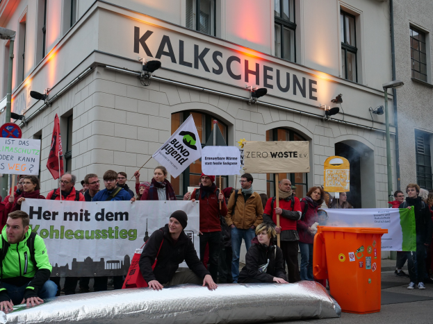 Menschen vor einem Gebäude mit Protestschildern und Transparenten, zwei Personen im Vordergrund sitzend und ein Müllcontainer rechts daneben.