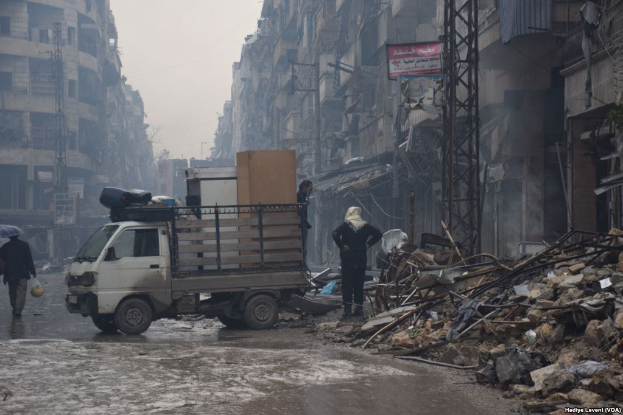 Ein Lastwagen fährt auf einer Straße neben einem Schutthaufen, mit Menschen, die auf der Straße gehen, einer hält einen Schirm, und Gebäuden, Polen und Schilder mit Text im Hintergrund bei klarem Himmel.