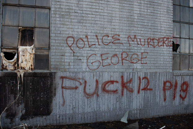 Ein Gebäude mit Graffiti, das 'Polizei Mord George Fuck 12 P19' lautet, mit Fenstern und diversen Gegenständen davor.