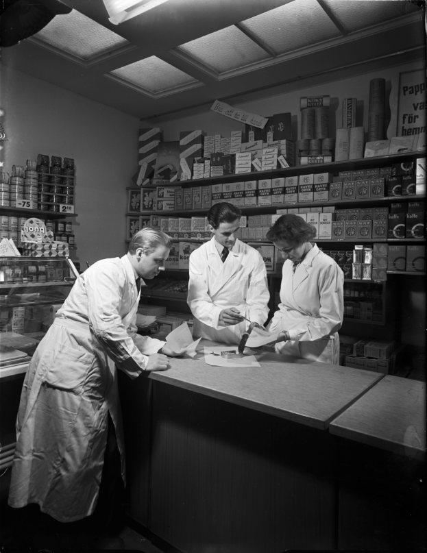 Drei Männer in weißen Kitteln stehen an einem Tresen in einer Apotheke mit Papieren und Gegenständen darauf, mit Regalen voller verschiedener Gegenstände im Hintergrund, in Schwarz-Weiß.