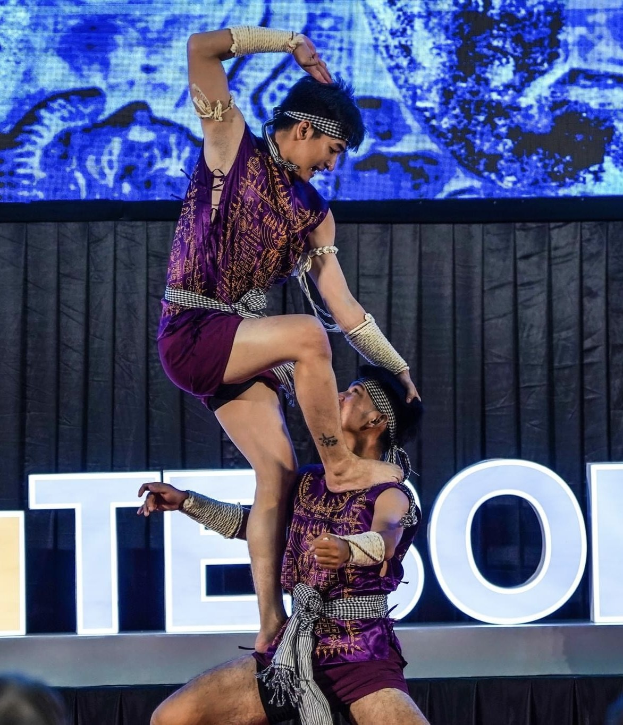 Zwei Akrobaten führen auf der Bühne eine Darbietung auf, der Mann hält die Frau in seinen Armen, während beide purpurne Kleider tragen, mit einem schwarzen Vorhang und einer Leinwand im Hintergrund und einem Publikum darunter.