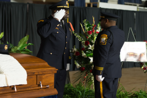 Zwei Polizeibeamte in Uniform stehen neben einem Sarg, der mit einem weißen Tuch bedeckt ist, mit Pflanzen, Blumen und Vorhängen im Hintergrund.