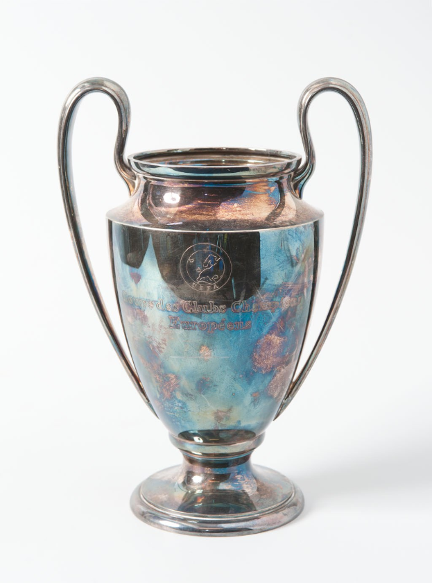 Der UEFA-Champions-League-Pokal, ein stählerner Becher mit zwei Griffen in Blau und Silber, mit der Inschrift "UEFA Champions League", vor einem weißen Hintergrund dargestellt.