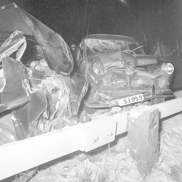 Schwarz-weiß-Foto eines verbeulten Autos mit eingedellter Motorhaube und zersplitterten Fenstern, das auf Gras neben einem Zaun mit dunklem Hintergrund geparkt ist.