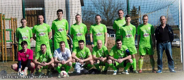 Eine Gruppe von Männern in grünen T-Shirts steht auf einem Fußballfeld, einige hocken, mit einem Ball vor einem Torpfosten, Bäume und Gebäude im Hintergrund.