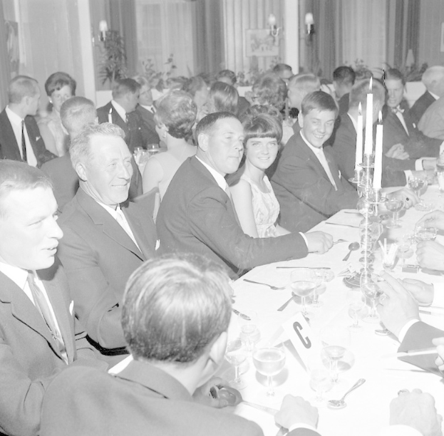Eine Gruppe von Menschen sitzt um einen langen Tisch, lacht und genießt die Gesellschaft des anderen, mit Gläsern, Besteck, Kerzen und anderen Gegenständen auf dem Tisch, vor einem Hintergrund aus Vorhängen und Wandlampen.