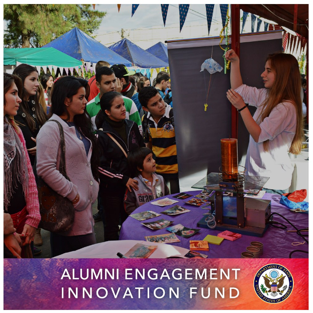 Gruppe von Menschen um einen Tisch versammelt mit einer Tafel, auf der "Alumni Engagement Innovation Fund" steht, mit Gegenständen auf dem Tisch und Zelten, Bäumen und einem klaren Himmel im Hintergrund.