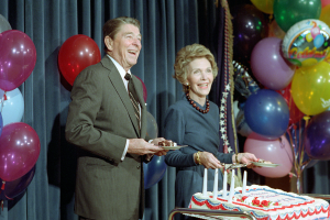Ein Mann und eine Frau stehen lachend neben einem Geburtstagskuchen mit Kerzen, halten Teller, mit bunten Ballons und einem Vorhang im Hintergrund.