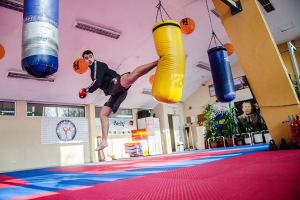 Ein Mann in der Luft, der einen Boxsack tritt in einer Sporthalle mit verschiedenen Geräten, Pflanzen und Fenstern.