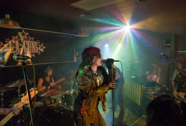Eine Frau in einer schwarzen Jacke singt in ein Mikrofon auf der Bühne, mit einem Mann in Sonnenbrille und Stirnbinde, der ein Instrument spielt, und einem Plakat mit 'LA Guns' im Hintergrund.