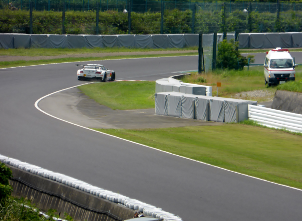 Ein Rennwagen fährt eine Rennstrecke entlang mit einem Polizeiwagen im Hintergrund, umgeben von Grünflächen und Absperrungen, mit Text unten im Bild.