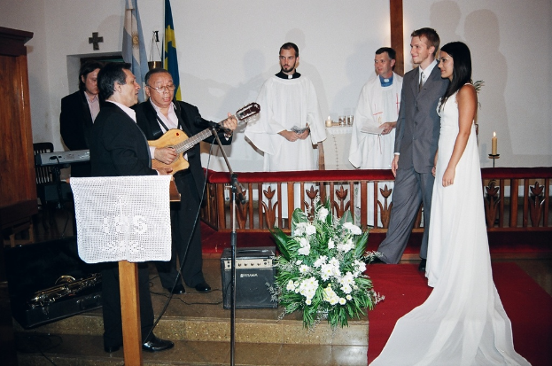 Eine Hochzeitszene in einem Raum mit zwei Pastoren, einem Brautpaar und drei Musikern; ein Podium mit Stoff vor den Musikern; ein Tisch mit Kerzen hinter den Pastoren; ein Kerzenhalter hinter dem Paar; ein Bukett, Lautsprecher, Taschen und roter Teppich auf dem Boden; sowie Flaggen, eine Wand, einen Schrank, einen Stuhl und ein Kreuz im Hintergrund.