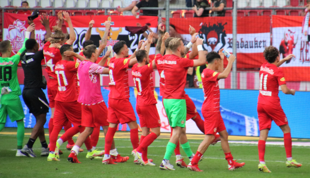 Gruppe von Fußballspielern, die ein Tor feiern, auf einem Feld, einige halten Flaschen oder klatschen, mit Bannern, Absperrungen und Stadion-Sitzplätzen im Hintergrund.