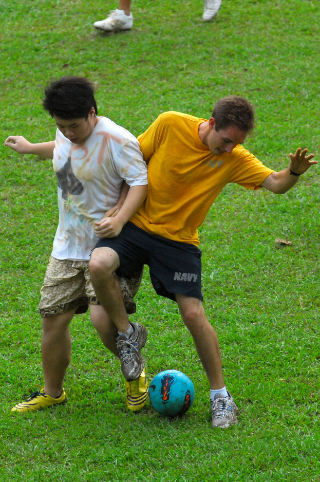 Zwei Männer spielen Fußball auf einem grünen Feld, einer trägt ein gelbes Shirt und schwarze Shorts, der andere ein weißes Shirt und braune Shorts, beide tragen Schuhe.