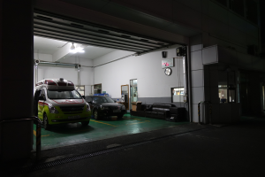 Ein Rettungswagen in einem Gebäude bei Nacht geparkt, mit sichtbaren Fahrzeugen, Sofas, einer Wanduhr, einer Texttafel, einer Tür, Rohren, Deckenleuchten und Metallstangen.