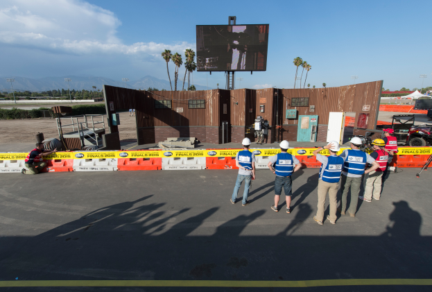 Eine Gruppe von Menschen in Helmen steht vor einem großen Bildschirm auf dem Las Vegas Motor Speedway, umgeben von Absperrungen, Fahrzeugen, Hütten, Bäumen, Laternenmasten und Bergen mit wolkenverhangenem Himmel im Hintergrund.