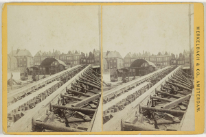 Schwarz-weiß-Foto einer Zugschiene mit Gebäuden im Hintergrund, Text 'Merkelbach & Co, Co. Amsterdam' rechts und hölzerne Gegenstände im Vordergrund verstreut.