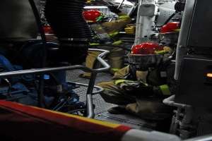 Feuerwehrleute in Helmen und Schutzausrüstung arbeiten an einem Feuerwehraufbau mit Rohren und einer Lampe im Hintergrund.