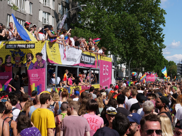 Eine große Menge marschiert auf einer Straße während des Berlin Pride 2019, viele tragen Kappen und Goggles, einige halten Fahnen, mit Bannern, Leuchtmasten, Bäumen und Gebäuden im Hintergrund unter einem bewölkten Himmel.