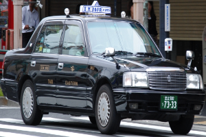 Ein schwarzes Taxi fährt die Straße entlang, vorne ein Radfahrer, im Hintergrund Gebäude, Pfosten und Schilder.