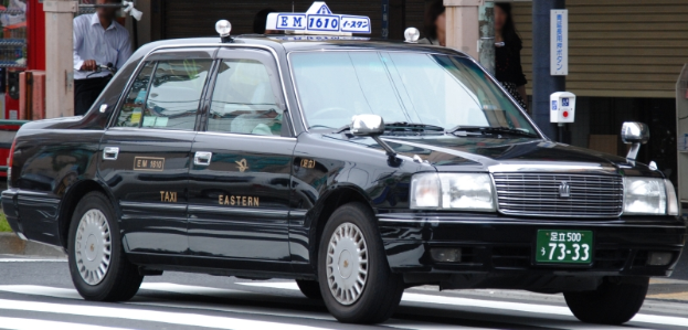Ein schwarzes Taxi fährt die Straße entlang, vorne ein Radfahrer, im Hintergrund Gebäude, Pfosten und Schilder.
