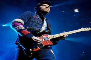 Ein Mann mit einer Mütze spielt Gitarre auf der Bühne bei einem Konzert, beleuchtet von hellen Scheinwerfern, mit einem freudigen und aufgeregten Gesichtsausdruck.