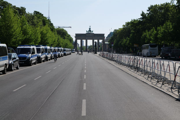Eine Reihe von Polizeiwagen, die auf einer Straße vor dem Reichstagsgebäude in Berlin, Deutschland, geparkt sind, mit Menschen auf Fahrrädern und auf der Straße, Barrieren, Bäumen und einem Bogen mit Statuen im Hintergrund.