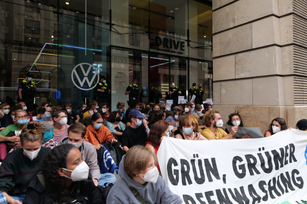 Große Gruppe maskierter Personen mit Spruchband vor einem Glasgebäude mit Logo demonstriert in Berlin, Deutschland und fordert den Abzug der Volkswagen AG.