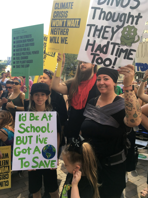 Eine Gruppe von Menschen mit Schildern, die gegen die Klimaveränderung protestieren, vor einer Menge, mit Bäumen, Gebäuden und einem klaren blauen Himmel im Hintergrund.
