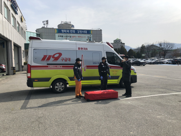 Ambulanz auf einem Parkplatz neben einem Geb├Ąude geparkt, mit drei Personen auf der Stra├če davor, gl├Ąserne Geb├Ąude auf der linken Seite, Fahrzeuge, B├Ąume, Berge und Himmel im Hintergrund.