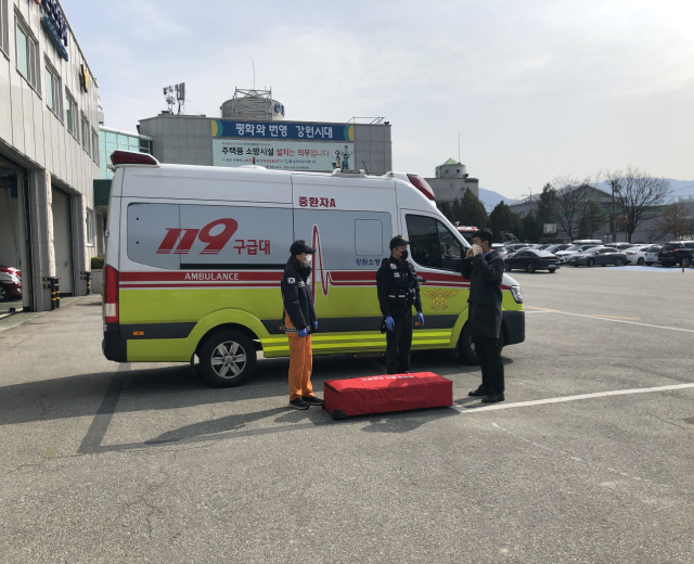 Ambulanz auf einem Parkplatz neben einem Geb├Ąude geparkt, mit drei Personen auf der Stra├če davor, gl├Ąserne Geb├Ąude auf der linken Seite, Fahrzeuge, B├Ąume, Berge und Himmel im Hintergrund.