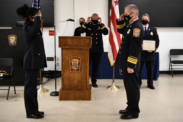 Gruppe uniformierter Personen in Masken vor einem Podium mit Mikrofon, umgeben von Flaggen und Möbeln, bei einer Vereidigung, bei der eine Person ein Zertifikat hält.