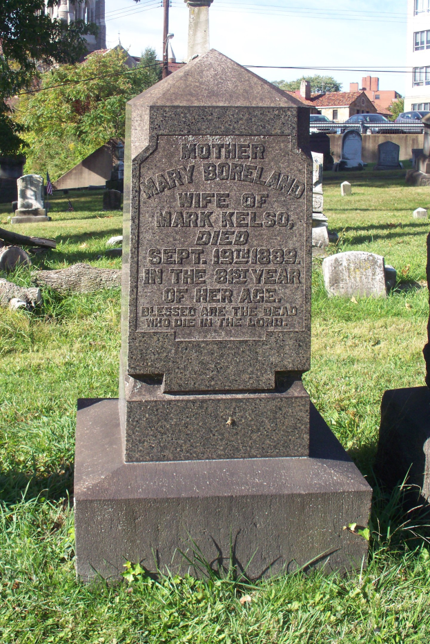 Grabstein inmitten eines grasbewachsenen Friedhofs mit Bäumen, Gebäuden, Strommasten und Drähten vor einem klaren blauen Himmel.
