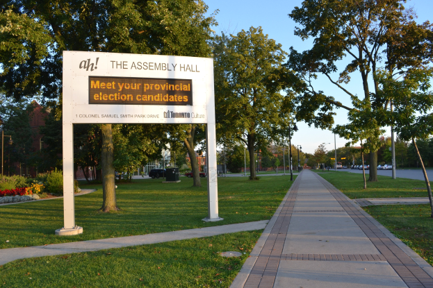 Plakat mit Text auf Gras neben Weg, Bäumen, Blumen, Straßenlaternen, Fahrzeugen, Gebäude und bewölktem Himmel; Text lautet "Die Versammlungshalle - Treffen Sie Ihre Kandidaten für die Landtagswahl."