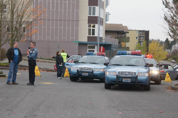 Autos auf einer Straße mit vier Personen in der Nähe, Gebäude mit Fenstern im Hintergrund, Bäume und Warn Dreiecke.