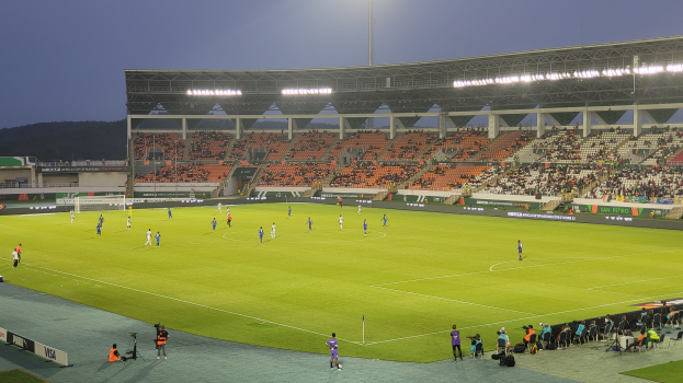 Ein Fussballspiel wird in einem Stadion mit Zuschauern auf Sitzen und Stehplätzen, einem Tor in der Mitte, einem Hügel und einem klaren blauen Himmel im Hintergrund und hellen Stadionlichtern gespielt.