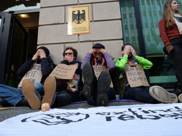 Eine Gruppe von Menschen sitzt vor einem Gebäude auf dem Boden, hält Schilder, mit einer Fahne und einem Apfel auf der Straße, und eine Frau steht rechts daneben und sieht besorgt aus.