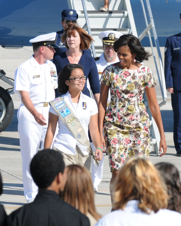 Michelle Obama und ihre Tochter Malia Obama gehen über das Rollfeld eines Flughafens, umgeben von uniformierten Personen, mit einem Fahrzeug und einer Treppe im Hintergrund.
