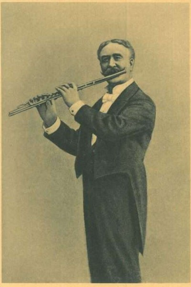Altes Schwarz-Weiß-Foto eines Mannes in einem Smoking, der auf der Bühne eine Flöte spielt und dabei die Augen geschlossen hat, mit zurückgekämmtem Haar und Bühnenbeleuchtung, die sein Gesicht erhellt.