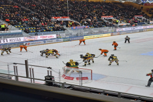 Gruppe von Menschen, die Hockey auf einer Indoor-Eisfläche mit einem Netz spielen, umgeben von einem Zaun, unter Stadionbeleuchtung, mit Zuschauern im Hintergrund und Schildern im Bild.