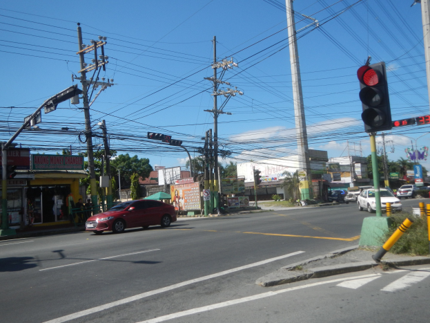 Ein rotes Auto fährt an einer Ampel auf einer Straße vorbei, die von Strommasten und Drähten gesäumt ist, mit Gebäuden, Bäumen und einem klaren blauen Himmel im Hintergrund.