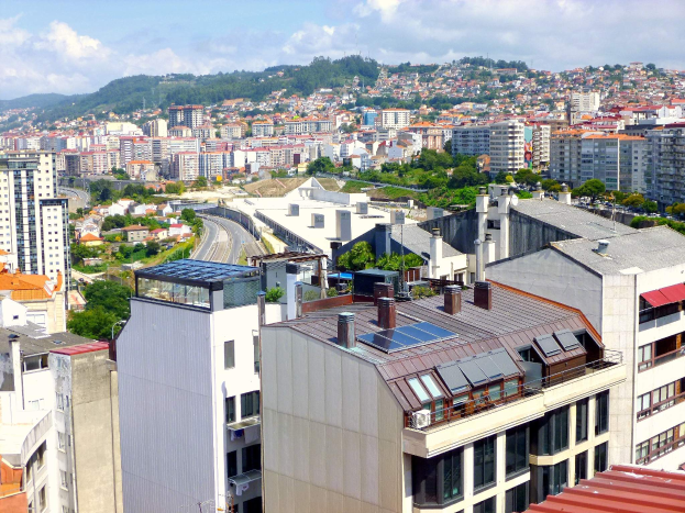 Eine Stadtansicht von einem Gebäudedach aus, mit zahlreichen Gebäuden, Bäumen, Straßen, Fahrzeugen und Bergen im Vordergrund, mit einem bewölkten Himmel im Hintergrund und Solarpanelen auf dem Dach eines Gebäudes.