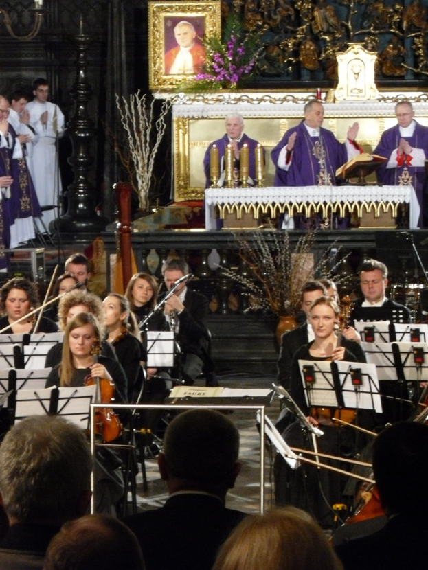 Eine Gruppe von Menschen, die Geigen spielen, mit Noten vor sich, während im Hintergrund drei Kirchenväter hinter einer Bibel stehen, mit einem von Schnitzereien umgebenen Fotoframe dahinter.