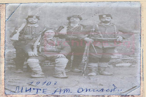 Ein Schwarz-Weiß-Foto einer Gruppe von Männern in Militäruniformen, einer sitzend mit einer Waffe in der Hand, mit dem Text "1910-1902 - munte am omisch" unten.