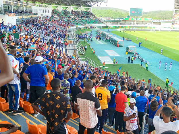 Eine große Menschenmenge versammelt sich vor einem eingezäunten Fußballfeld unter einem klaren blauen Himmel, mit Hügeln im Hintergrund, einige sitzen auf Stühlen und andere stehen, und warten auf ein Spiel.