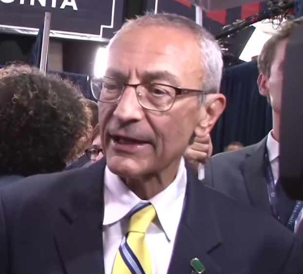 Ein Mann im Anzug und Krawatte, der eine Brille trägt, spricht zu Reportern mit einem Mikrofonständer vor sich, umgeben von einer Gruppe von Menschen, mit Lichtern und einer Tafel im Hintergrund.